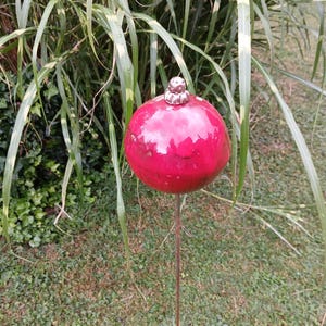Könnte beinhalten: Ein glänzendes, kugelförmiges rotes Gartenornament mit einer metallisch silberfarbenen Kappe an der Oberseite. Das runde Objekt ist auf einem dünnen, geraden Metallstab montiert, der zum Einstecken in den Boden gedacht ist und einen Akzent setzt.