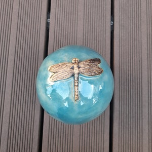 Op de afbeelding: Een rond, turquoise keramisch decoratief object met een bronzen libellenontwerp. De libel is gecentreerd op de bovenkant van de bol. Het object ligt op een bruin houten oppervlak.