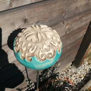 Peut inclure: Un piquet de jardin décoratif avec une sphère beige à motifs floraux et une base turquoise. La sphère est montée sur une fine tige métallique sombre. Le piquet est placé dans un parterre de jardin avec de petits cailloux et de la verdure. Diamètre: environ 15 cm.