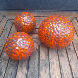 May include: Three orange ceramic spheres with a textured surface and a glossy finish. The spheres vary in size and are arranged on a wooden surface. The color is a vibrant orange with darker brown accents.
