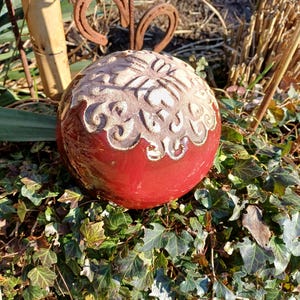 Op de afbeelding: Een rode keramische tuinsfeer met een gedetailleerd, crèmekleurig bloemmotief. De bol staat te midden van groene klimop en wordt bekroond met een decoratief, roestig metalen accent. Een uniek buitentuindecoratie.
