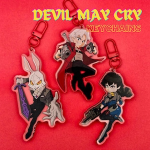 May include: Three acrylic keychains featuring characters from "Devil May Cry." The keychains have red clasps and depict a white-haired character in a red coat, a character with a gun, and a rabbit-like character with a sword. The text "DEVIL MAY CRY KEYCHAINS" is at the top.