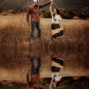 Può includere: Una coppia balla in un campo di erba alta. L'uomo indossa una camicia marrone, jeans blu e un cappello da baseball. La donna indossa una camicia bianca, una lunga gonna marrone e nera e ha i capelli raccolti. La coppia si riflette in una pozza d'acqua.