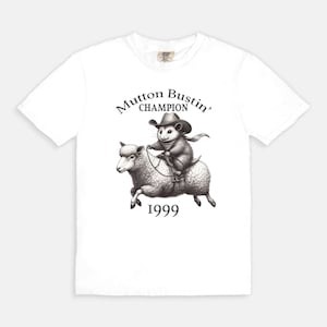 May include: White t-shirt with a black and white illustration of a possum riding a sheep. The text "Mutton Bustin' Champion 1999" is printed on the shirt.