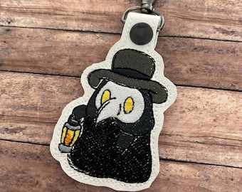 Può includere: Portachiavi ricamato con un dottore della peste dei cartoni animati con cappello nero, maschera e mantello, che tiene una lanterna. Il portachiavi ha un retro bianco e una chiusura color argento.