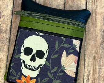 Può includere: Piccola pochette rettangolare con parte superiore blu scuro e cerniera verde. La parte anteriore presenta un disegno a teschio su sfondo floreale blu navy. La pochette è larga circa 13 cm e alta 10 cm.