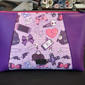Peut inclure: Pochette zippée violette avec intérieur rose. Le panneau central présente un motif répété de consoles de jeux, de crânes, de nœuds et de chauves-souris sur un fond quadrillé. Les côtés sont violets unis.