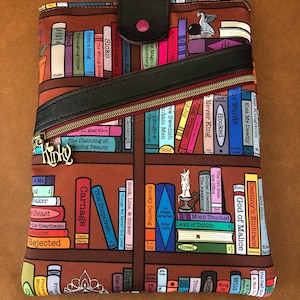 May include: A brown and black book sleeve with a zipper closure and a bookshelf pattern featuring the titles of various books. The sleeve has a black leather flap with a pink button.