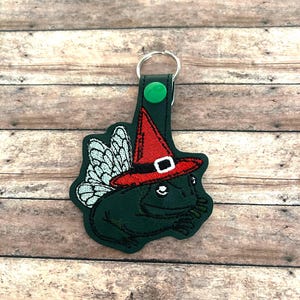 Pode incluir: Um chaveiro em forma de sapo verde escuro com um chapéu de bruxa vermelho e asas brancas. O chapéu tem uma faixa e uma fivela pretas. O chaveiro tem um botão de pressão verde e um porta-chaves prateado.