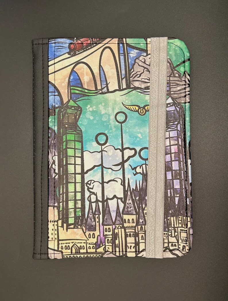 May include: A black passport holder featuring a colourful illustration of Hogwarts castle, the Hogwarts Express, and the Golden Snitch. The holder has a silver elastic band to keep it closed.
