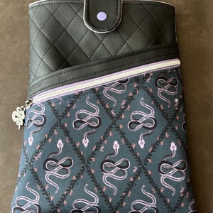 May include: Black quilted zippered pouch with a purple button closure. The pouch is lined with a fabric featuring a repeating pattern of pink snakes on a dark green background.