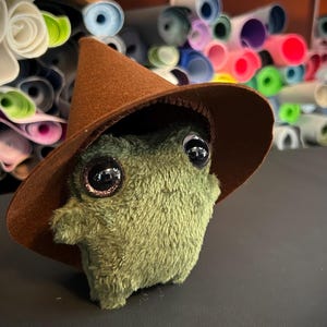 Puede incluir: Una criatura verde de peluche con grandes ojos brillantes y un sombrero de bruja marrón. El peluche tiene una textura suave y pequeños brazos. En el fondo, se ven rollos de tela colorida.