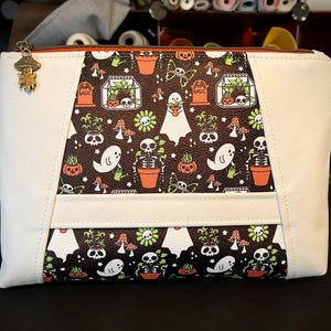 Peut inclure: Une pochette zippée blanche et marron avec un motif sur le thème d'Halloween. Le motif comprend des fantômes, des squelettes, des citrouilles et des plantes en pot. La tirette de la fermeture éclair est un charme en forme de champignon.