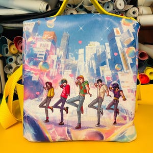 Puede incluir: Una mochila cuadrada azul y blanca con asa y correa amarillas. La mochila presenta una colorida ilustración de un paisaje urbano con cinco figuras bailando. El diseño incluye corazones y formas esféricas.