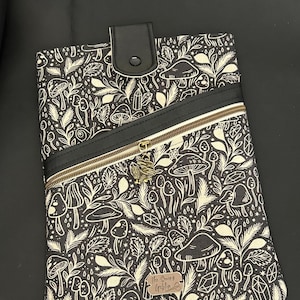 May include: Tablet case with a black and white mushroom and foliage pattern. It has a black zip, a black leather flap with a snap closure, and a small metal charm. The text "The Swiny Goblin" is on a small leather tag.