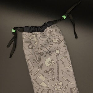 Puede incluir: Una bolsa de cordón gris con un cordón y un ribete negros. La bolsa está impresa con un patrón de varias ilustraciones de temática fantástica, incluyendo espadas, escudos, pociones y una calavera.