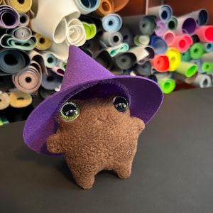 Puede incluir: Una criatura de peluche marrón con grandes ojos brillantes, que lleva un sombrero de bruja de fieltro morado. El juguete hecho a mano tiene un cuerpo simple y redondeado y pequeños brazos y piernas. El fondo muestra rollos de fieltro de colores.