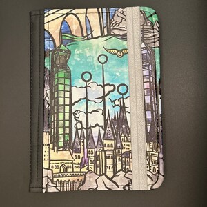 May include: A colourful journal with a fantasy castle design. The cover features a detailed illustration of a castle, towers, and a bridge, with a grey elastic band closure. The design includes a golden snitch and quidditch hoops.