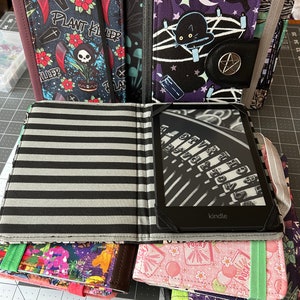 May include: A stack of six colourful fabric book covers with different designs. The top cover is black and white striped with a Kindle e-reader inside. The second cover is pink with a white and pink floral pattern. The third cover is green with a purple and green floral pattern. The fourth cover is brown with a multi-coloured floral pattern. The fifth cover is black with a purple and black floral pattern with a black cat and a pentagram. The sixth cover is black with a skull and crossbones and the text "Plant Killer".