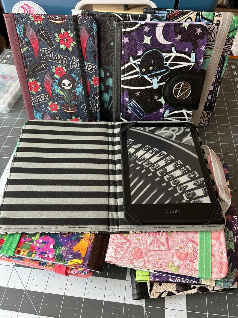 May include: A stack of six colourful fabric book covers with different designs. The top cover is black and white striped with a Kindle e-reader inside. The second cover is pink with a white and pink floral pattern. The third cover is green with a purple and green floral pattern. The fourth cover is brown with a multi-coloured floral pattern. The fifth cover is black with a purple and black floral pattern with a black cat and a pentagram. The sixth cover is black with a skull and crossbones and the text "Plant Killer".