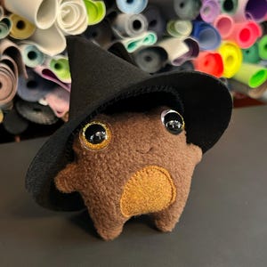 May include: A brown plush toy frog wearing a black witch hat with gold trim. The frog has large, expressive eyes and a tan belly. The background features rolls of colorful fabric.