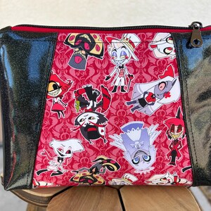 May include: A black and red zippered pouch with a glittery finish. The pouch features a repeating pattern of cartoon characters from the animated series "Hazbin Hotel".