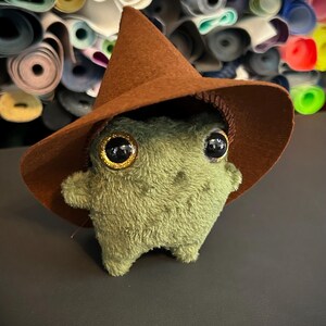 Puede incluir: Un peluche verde con grandes ojos con borde dorado y un sombrero de bruja de fieltro marrón. El juguete hecho a mano tiene brazos y piernas pequeños, sobre un fondo de rollos de tela coloridos.
