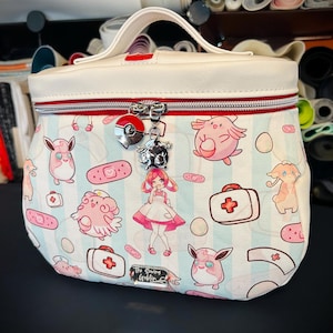May include: A white cosmetic bag with a red zipper and handle, featuring a cartoon print of pink characters, bandages, and medical kits on a light blue striped background. Charms hang from the zipper. The Sewing Goblin is printed on the bottom.