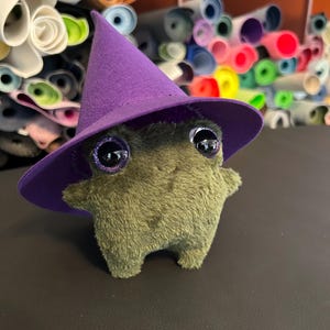 Puede incluir: Un peluche de rana verde con un sombrero de bruja morado y ojos grandes y brillantes. El fondo muestra rollos de tela colorida, lo que sugiere un tema de artesanía o costura.
