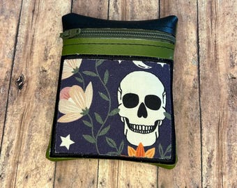 Può includere: Piccola pochette rettangolare con parte superiore blu scuro e cerniera verde. La sezione inferiore presenta un motivo floreale e teschio su sfondo blu scuro. La pochette è rifinita in verde e ha un retro nero.