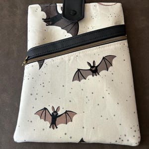 May include: A cream-coloured pouch with a bat pattern and a black leather accent. The pouch features a black zip, a black leather flap with a snap closure, and a repeating pattern of bats and stars.