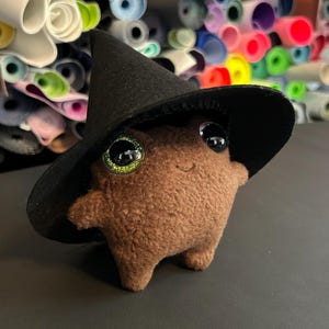 May include: A brown plush creature wearing a black witch hat with large, glittery green and black eyes. The plush toy has a smiling face and small arms and legs. The background is blurred with rolls of colorful fabric.