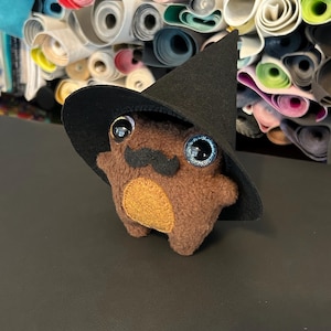 Puede incluir: Un peluche de rana marrón con grandes ojos azules brillantes, un bigote negro y una mancha amarilla en el vientre. La rana lleva un sombrero de bruja negro. El fondo presenta rollos de tela colorida.