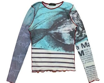 May include: A long-sleeved, blue and white striped shirt with a fish graphic and text on the sleeves. The text on the sleeves reads "100% le" and "a Ma M EME oh anh reus, lo cite o de sorts Marseille de THotobu guidée des Depart".