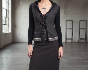 May include: A dark grey hooded jacket with black sleeves and a matching skirt. The jacket has a unique button closure with brown ties and a decorative trim. The skirt features a subtle geometric pattern, creating a coordinated ensemble.