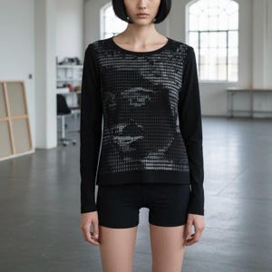 Peut inclure: Haut noir à manches longues avec un motif graphique en niveaux de gris sur le devant, associé à un short noir. Le motif semble être une image pixélisée. La personne est dans une pièce lumineuse avec de grandes fenêtres.