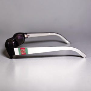 May include: Black and white sunglasses featuring a prominent Gucci logo on the temple. The side arm displays a horizontal green and red stripe detail with a gold-toned circular emblem containing the interlocking G logo on a white background.