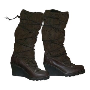 Puede incluir: Un par de botas de cuña marrones. Las botas tienen una parte superior de nailon acolchado con un diseño de cordones y una sección inferior de cuero. El tacón de cuña es negro.