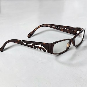 May include: Brown tortoiseshell eyeglasses with rectangular frames. The Prada logo is displayed in white on the temples. The glasses have a classic design and are suitable for everyday wear.