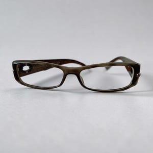 May include: Rectangular spectacles with a translucent dark brown frame. The temples feature a distinct oval cutout detail near the hinges, showcasing a dark brown pattern. The frames have a smooth, polished finish and clear lenses.