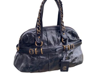 Pode incluir: Uma bolsa de mão de couro preto com ferragens douradas e uma alça de corrente. A bolsa tem formato de cúpula e apresenta várias fivelas e correias.