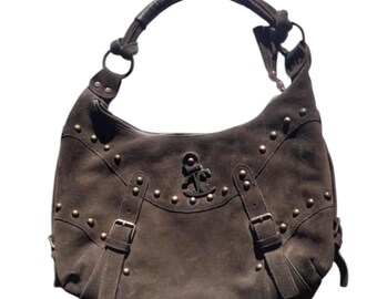 May include: Brown suede shoulder bag with a curved shape and a dark brown leather handle. The bag features gold-coloured studs and buckles, with an anchor emblem on the front. A stylish accessory.