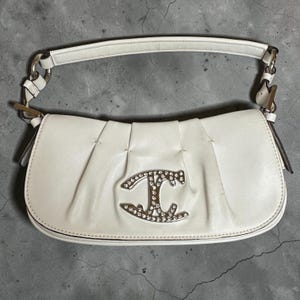 Puede incluir: Un bolso de hombro blanco con frente plisado y un emblema de metal plateado con cristales transparentes incrustados. El bolso tiene una sola asa superior sujeta con herrajes plateados y una silueta curva y estructurada con bordes lisos.