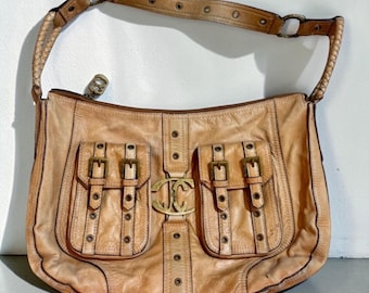 May include: A tan leather shoulder bag featuring two front pockets with brass-toned buckles and grommets. A central vertical leather strip displays a large, ornate gold-toned metal logo. The bag includes a braided leather strap and a zipper closure.
