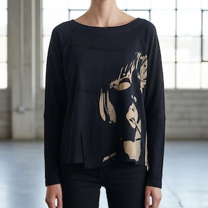 May include: Black long-sleeve shirt with a gold-colored abstract design on the front. The shirt has a scoop neck and a relaxed fit. The design appears to be a stylized face or figure. The shirt is paired with black pants.
