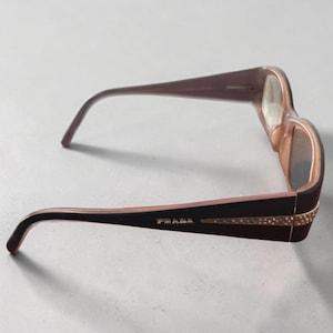 May include: Brown Prada eyeglasses with a rectangular frame. The temples are dark brown with the brand name in gold lettering and embellished with small, sparkling details. The lenses are a light brown tint.