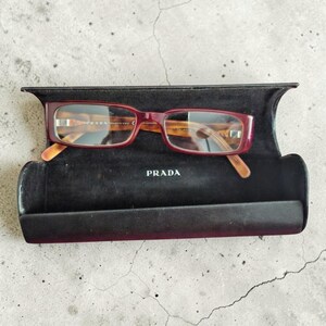Peut inclure: Lunettes rectangulaires Prada avec une monture rouge foncé et des branches à motif écaille de tortue marron, reposant dans un étui rigide doublé de velours noir avec le logo Prada imprimé en blanc sur la base intérieure.