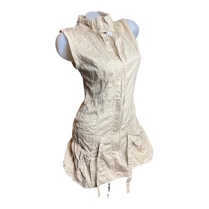 May include: A beige sleeveless dress with a ruffled collar, a button fastening, and a cinched waist. The dress has a tiered skirt with pockets and a drawstring detail.