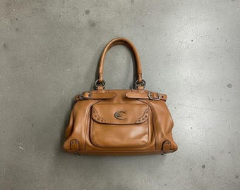 May include: A tan leather handbag with a top handle and a front pocket. The bag has silver-tone hardware, including buckles and rivets. The front pocket features a decorative emblem. The bag is sitting on a grey surface.