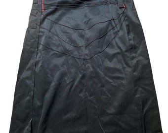 May include: A black knee-length skirt featuring a series of thin, horizontal black cords draped across the upper front section. The skirt includes red fabric accents visible along the side seam and at the waist, with three square stitched tabs.
